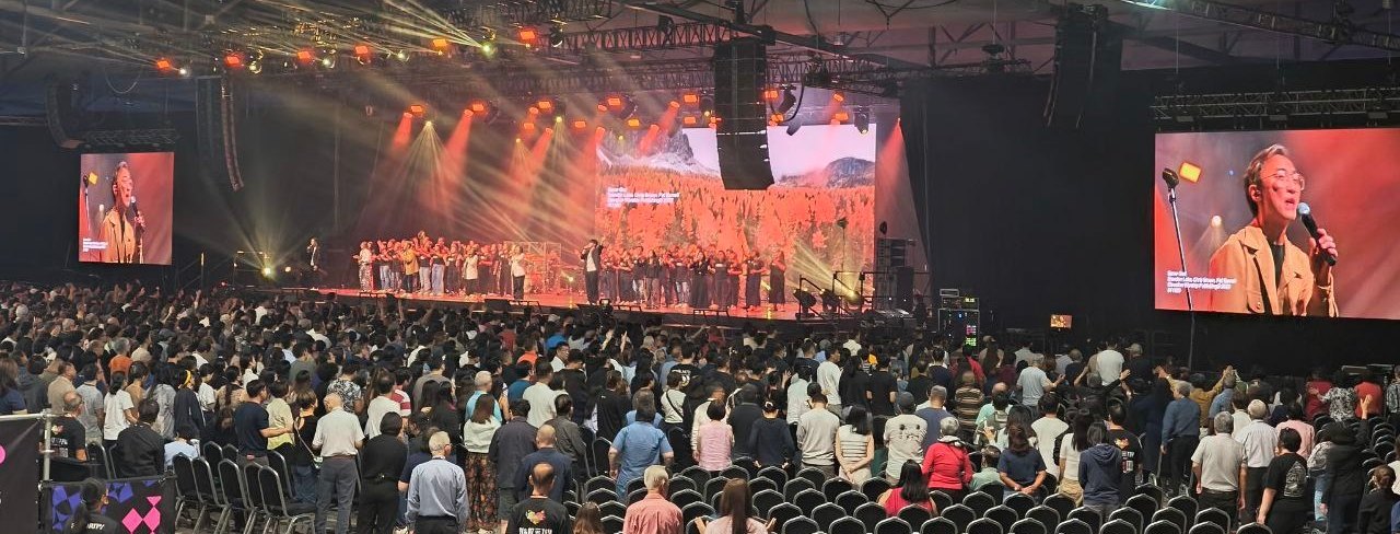 A full exhibition hall standing for a live performance, with side screens and warm stage lighting