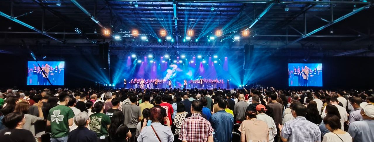 A packed exhibition hall facing a lit concert stage, with side screens for the rows at the back