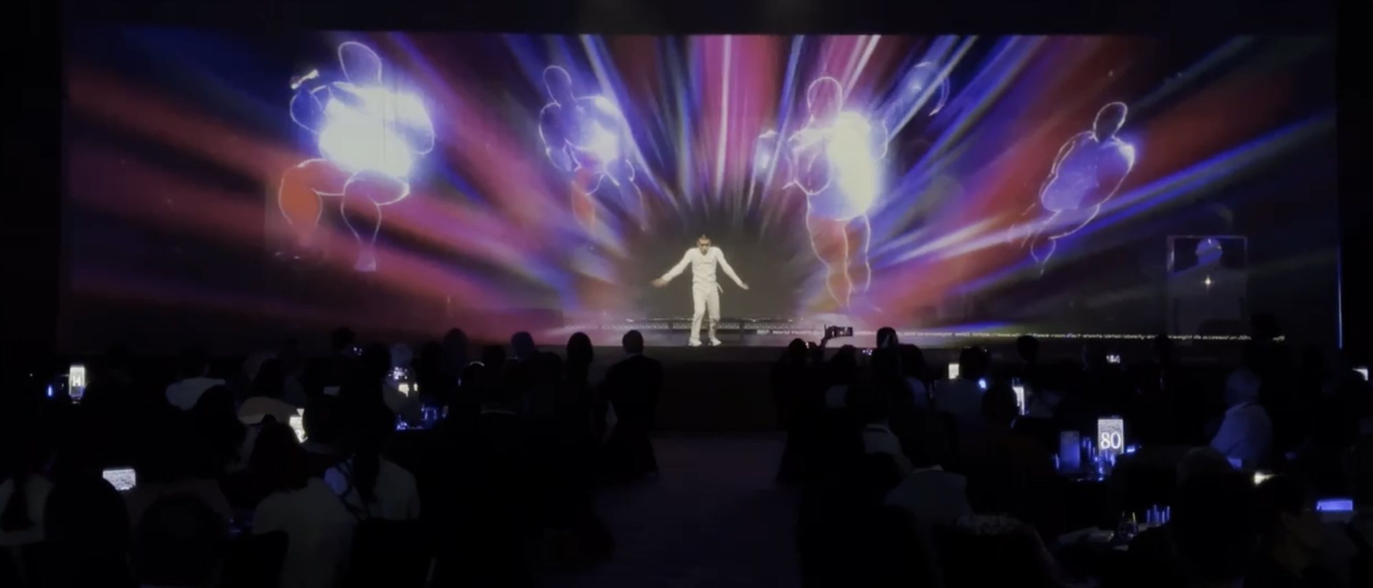 A performer on stage surrounded by holographic figures and light, in front of a seated audience