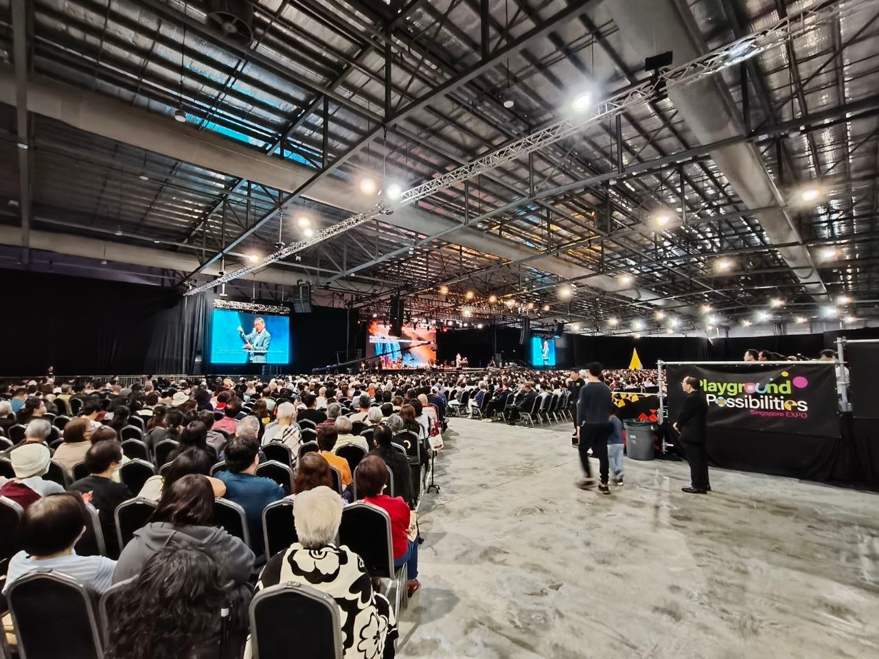 A full exhibition hall seated for a live event, with side screens and a wide aisle