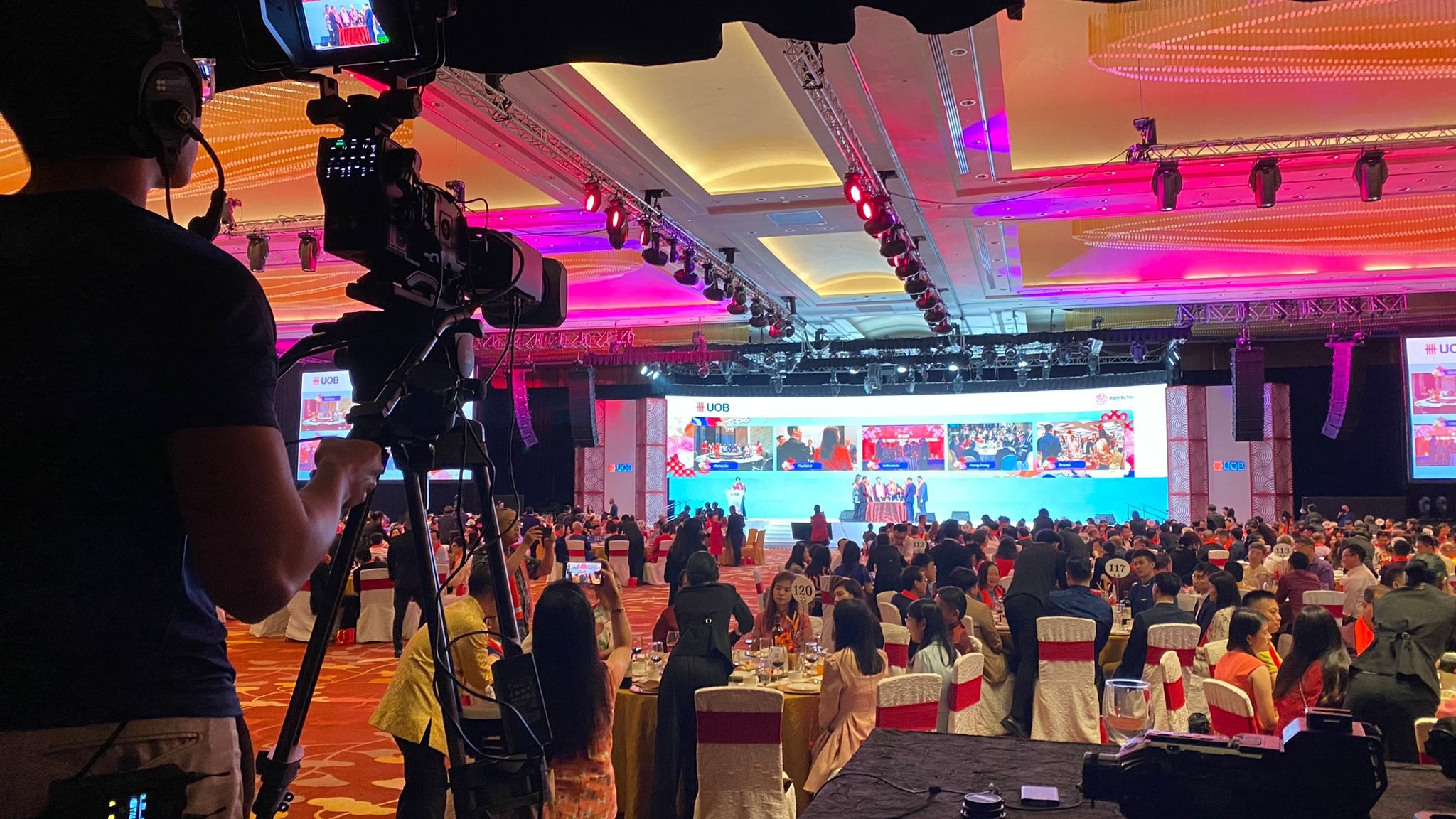 A camera operator filming a packed ballroom celebration, with the stage on screen