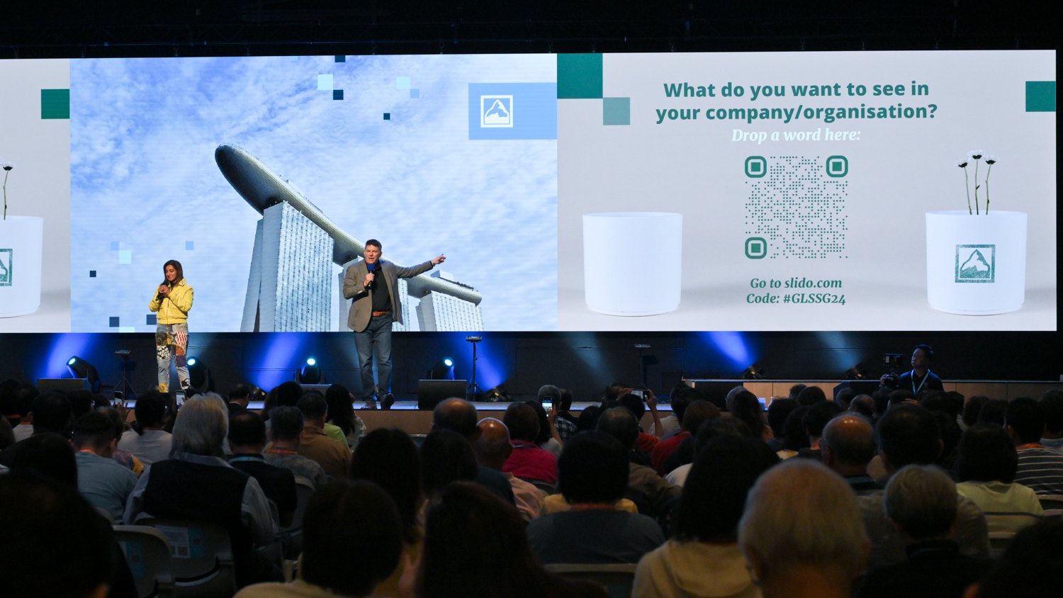 A keynote speaker on stage in front of a wide LED wall, with the audience in the foreground